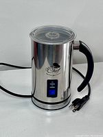 Front and side view of stainless steel electric milk frother showing handle, brand logo, control panel, cord and plug.