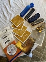 Overview photo showing several packs of new guitar strings, a silver harmonica case with a blue cover in background, harmonica, and brass tuning knobs in plastic bag.
