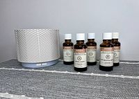 White Saje Aroma Nook diffuser next to five 1-oz bottles of Aveda essential oil blends on a gray mat