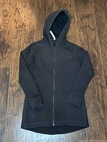 Front view of black Lululemon jacket laid flat on wooden floor, showing full zipper and hood.