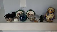Overview shelf photo showing stone bird figurines, embroidered trinket boxes, glass paperweights, green bird ceramic figurine, and the largest Russian nesting doll figurine.