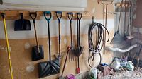 Full view of wall with multiple garden tools including shovels, rake, broom, brushes, dustpan, and coiled garden hose.