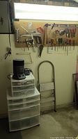 Photo showing pegboard with various hand tools hung including saws, wrenches, clamps, screwdrivers, hammer, and white plastic storage drawers below with a black motor or device on top.