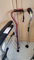 Photo of red and silver walking canes alongside an aluminum walker with black padded armrests.
