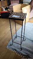 Full view of the wrought iron plant stand showing its height, base, and square tiled top.