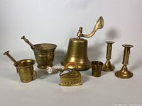 Group photo showing all seven brass items including the wall bell, mortar and pestles, candleholders, cup, and iron decoration.