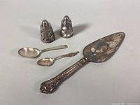View of two sterling silver spoons, two etched salt and pepper shakers, and a cake cutter with a dented blade and detailed handle.
