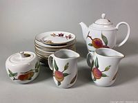 Photo of porcelain tea set components including teapot, sugar bowl, two creamers, and stack of dessert plates showing fruit designs and gold trim.