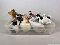 Clear view of assortment of ceramic and porcelain cow creamers and jugs tightly packed in plastic container.