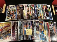 Photo of a collection of approximately 50 comic books arranged in two rows, showing a mix of titles including Justice League, Batman, and X-Men. Comics are individually bagged and boarded.