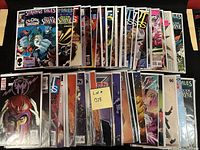 Photo shows two rows of assorted comic books stacked on a black surface, each comic bagged and boarded for protection. Titles visible include Strange Tales, Son of M, Doctor Strange, and others from both Marvel and DC universes.
