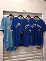 Four Blue Jays baseball jerseys hanging on hangers, three blue and one light blue.