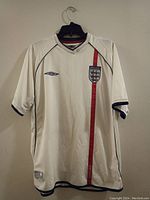 Front view of white England soccer jersey displaying red vertical stripe, Umbro logo, and England team crest.
