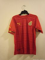 Front view of the red Ghana Football Association football jersey with Puma logo and team crest, displayed on a hanger against a beige wall.