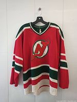 Front view of a red New Jersey Devils hockey jersey hanging on a hanger against a white background. The jersey has prominent team logo and colored stripes.