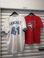 Back view of Sanchez #41 white baseball jersey and front of red Blue Jays T-shirt, both hanging on black hangers.