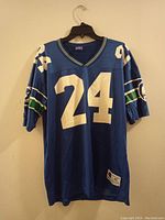 Front view of blue Seattle Seahawks jersey with number 24, showing Champion branding and sewn tags, and team colors on sleeves and collar.