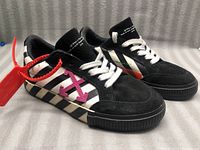 Pair of black Off-White Vulcanized Low sneakers with white diagonal stripes and purple arrow detail, white laces, and black vulcanized sole.