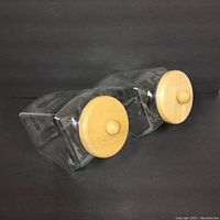 Angled side view of two clear rectangular glass canisters with round wooden lids and knob handles, lying on their side on a black background.