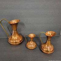 Set of three copper vases shown upright with brass handles, different heights displayed against a grey background.
