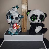 Both plush toys seated with ruler showing 5.5 to 6 inches height. Gia the giraffe on left with gray and white fur, turquoise accents and large glitter eyes. Bamboo panda on right with black and white fur, large green glitter eyes and black ears with green inner color.