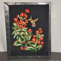Full view of the needlepoint artwork featuring orange flowers and a hummingbird on black fabric stretched and framed