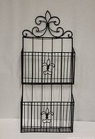 Front view showing two-tier black wire hanging magazine rack with Flor de Les fleur-de-lis decorative elements