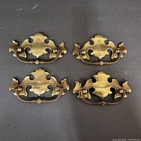 Four ornate vintage brass drawer pulls laid out on dark surface, showing front and handle design