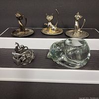 Set of five cat-themed holders on two-tiered white display stand with black background. Top tier holds three pewter cat candle holders with round trays. Bottom tier holds pewter cat ring holder with open design and glass curled cat candle holder.
