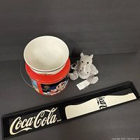 Photo showing all items in the lot: Coca Cola bar mat, red tin can, white bowl, and gray plush rhinoceros