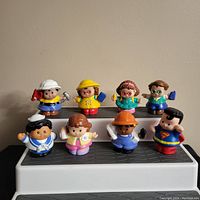Eight Fisher Price Little People plastic figures arranged on a white and black surface, front view showing the varied characters including a Superman figure, construction worker, and others in colorful outfits.