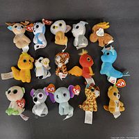 Top view of 15 small Ty Beanie Babies and plush toys, showing a variety of animals including deer, blue dog, owl, dove, moose, lion, panda, tiger, cardinal, unicorn, monkey, koala, elephant, giraffe, and a small deer or antelope, each with Ty heart-shaped hang tag.