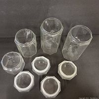 Four clear glass jars with octagonal shape and matching white screw-on lids displayed on a black surface, seen from above showing lid details and clear transparency.