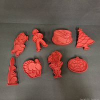 Eight red plastic cookie cutters arranged on a black surface in shapes including pig, scarecrow, slice of cake, Christmas tree, rabbit, turkey, bouquet of flowers, and jack-o'-lantern pumpkin.