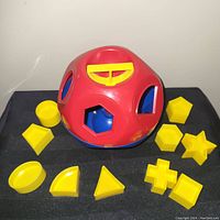 Red plastic shape sorter toy with yellow shape pieces arranged around it on a black surface, showing condition and detail of holes and pieces.
