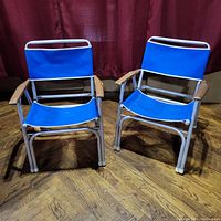 Front view of two vintage aluminum folding lawn chairs with blue fabric seats and wooden armrests, placed on wood floor in front of red curtain.