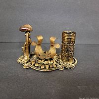 Front and angled view of vintage gold tone metal lipstick holder showing floral filigree base, central lipstick holder, and cylindrical container with perforated design.
