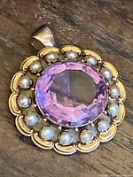 Front view of circular 14K yellow gold pendant showing large faceted purple amethyst center surrounded by cultured pearls on wooden surface.