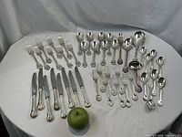 Full layout of all 44 silver-plate pieces on white cloth with green apple for scale