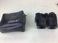 Black compact binoculars with central focusing wheel and textured grip details, placed next to a black zippered carrying case with Jason branding.