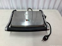 Top image of the Breville panini press showing stainless steel lid and black handle, plugged in with visible cord.