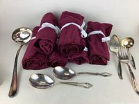 Rolled burgundy velvet storage bags with white ribbons, accompanied by various silverplated flatware including ladle, large spoons, and fork.