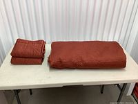 Photo showing folded copper colored quilted bedspread and two folded pillow shams on a table