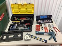 Black plastic toolbox filled with various hand tools. Yellow lid of toolbox is open. Socket wrench set in blue case and manual caulk gun placed next to toolbox. Tape measure and carpenter's level placed in front. Multiple hand tools like pliers and wrenches arranged on surface.