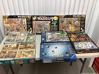 Nine assorted puzzles displayed on table, mostly Wasgij series including Wasgij 3, 9, 24 and Ravensburger puzzle