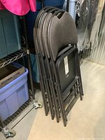 Four gray folding chairs stacked together, showing padded backs with top handles and black metal frames.