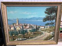 Framed original signed seascape painting showing a coastal village scene with trees, a winding path, water, and mountains in the background. The image highlights the painting's colors, brushwork, and frame detail.