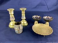 Photo of four vintage brass pieces: two candlesticks, one trinket bowl, and one toothpick holder, arranged on a dark cloth background showing patina and wear.