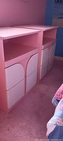 Two matching pink and white night stands shown against pink carpet and blue-pink walls, each features an open top shelf and two cabinet doors with arch design.