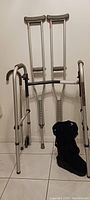 Photo showing a pair of aluminum crutches, a folding walker, and a black orthopedic fracture boot leaning against a wall on tiled floor.